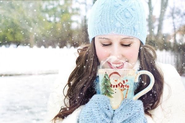 Woman in a winter cap and mittens, smelling coffee from a holiday mug.
